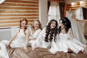 Four women are sitting on a bed, smiling and laughing together. They are all wearing white dresses, and one of them is wearing a tiara. Scene is happy and joyful photo