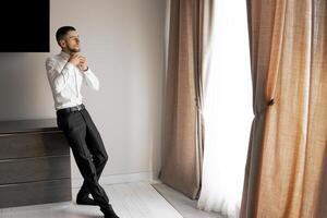 A man in a suit is standing in front of a window with curtains. He is looking out the window, possibly thinking about something photo
