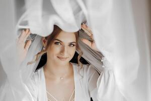 A woman is wearing a white dress and is smiling. She is standing in front of a curtain, with her head partially covered by it photo