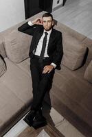 A man in a suit and tie is sitting on a couch. He is wearing a black suit and a white shirt photo