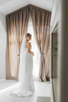 A bride stands in front of a window with a white dress and veil. The curtains are drawn, and the room is dimly lit. The bride appears to be waiting for someone, possibly her groom, to arrive photo