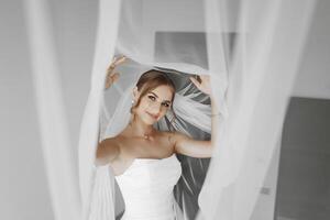 A bride is wearing a white dress and veil, and is standing in front of a curtain. The image has a romantic and intimate mood, as the bride is posing for a picture in front of the curtain photo