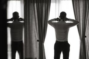 A man in a suit is standing in front of a window, adjusting his tie. The image has a moody and contemplative feel, as the man is lost in thought while getting ready for an event photo