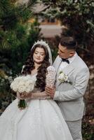A bride and groom are posing for a picture in front of a tree. The bride is wearing a white dress and holding a bouquet of white flowers. Scene is happy and romantic photo