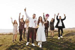 A group of people are celebrating a wedding, with the bride and groom in the center. The bride is wearing a white dress and the groom is wearing a suit. The group is standing in a field photo