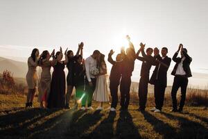A group of people are posing for a picture on a grassy hill. The bride and groom are in the center of the group, with their arms around each other. The rest of the group is standing behind them photo