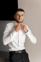 A man in a white shirt and black pants is standing in front of a wall. He is adjusting his shirt and looking at the camera. The image has a casual and relaxed mood photo