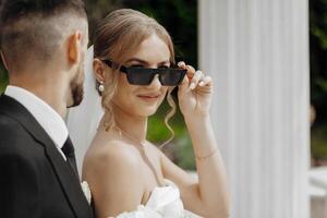A bride and groom are posing for a picture with sunglasses on. The bride is wearing a white dress and the groom is wearing a black suit. The sunglasses add a cool and stylish touch to the photo