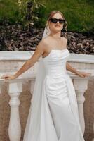 A woman in a white dress is standing on a balcony, wearing sunglasses and a white veil. She is smiling and she is enjoying the view photo