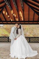 A bride and groom are posing for a picture in front of a wooden structure. The bride is wearing a long white dress and the groom is wearing a suit. Scene is happy and romantic photo