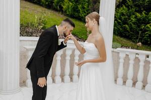 A bride and groom are standing on a white marble balcony, the bride is wearing a white dress and the groom is wearing a black suit photo