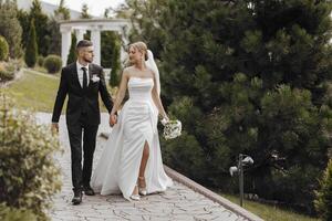 A bride and groom are walking down a path in a park. The bride is wearing a white dress and the groom is wearing a suit. They are holding hands and appear to be happy and in love photo