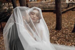 A bride and groom are embracing under a white veil. The bride is wearing a white dress and the groom is wearing a black suit. The scene is set in a forest, with trees in the background photo