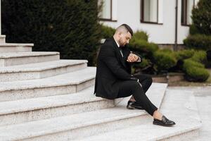 A man in a suit is sitting on a set of stairs. He is wearing a watch and he is checking the time. Concept of formality and professionalism, as the man is dressed in a suit and tie photo