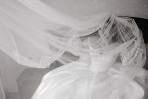 A bride is getting ready for her wedding and is wearing a white dress. The dress is flowing and has a veil on it. The bride is standing in front of a bed, and the bed is covered with a white sheet photo
