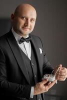 A man in a suit holding a box. The man is smiling and wearing a bow tie. The box is silver and has a shiny appearance photo