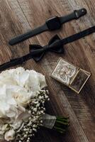 A bouquet of white flowers sits on a wooden table next to a watch and a tie. The flowers are arranged in a vase and the tie is folded photo