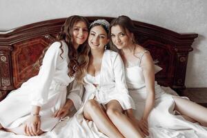 Three women are sitting on a bed, all wearing white dresses. They are smiling and seem to be enjoying each other's company. Scene is lighthearted and friendly photo