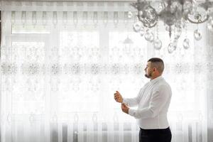 A man in a white shirt is standing in front of a window, adjusting his tie. The room is decorated with a chandelier and has white curtains. The man is in a hurry photo