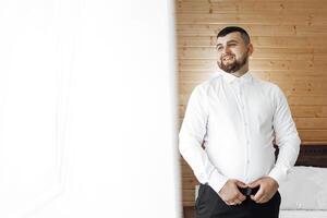 A man in a white shirt and black pants is standing in front of a window. He is smiling and looking out the window photo