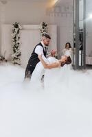 A bride and groom are dancing in the air in front of a foggy background. The bride is wearing a white dress and the groom is wearing a black vest photo