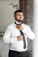 A man is wearing a black tie and smiling. He is standing in front of a window. The image has a light and cheerful mood photo