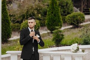 A man in a suit is getting ready for a wedding. He is adjusting his tie and looking at the camera. The scene takes place in a park with a white fence in the background photo