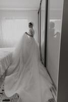 A woman in a wedding dress is standing in front of a mirror. The dress is long and flowing, and the woman is holding a veil. The image has a romantic and elegant mood photo