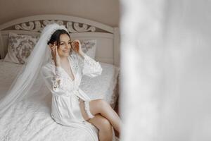 A bride is sitting on a bed with a veil on her head. She is wearing a white gown and is looking at the camera photo