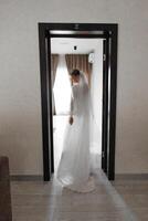 A bride is standing in front of a door, wearing a white dress and veil. The image has a moody and dramatic feel, as the bride is the only person visible in the frame. The door appears to be closed photo
