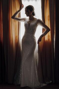 A woman in a long white dress is standing in front of a window. She is wearing a veil and has her arms outstretched photo