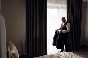 A man in a suit is standing in front of a window, getting dressed. The room is dimly lit, and the curtains are drawn, creating a sense of privacy and intimacy photo