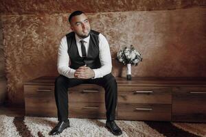 A man in a suit and tie sits on a wooden table with a vase of flowers in front of him photo