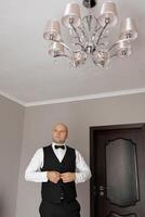 A man in a suit and vest stands in front of a chandelier. He is adjusting his tie and looking up at the ceiling. Scene is formal and elegant, as the man is dressed in a suit and vest photo