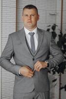 A man in a suit and tie is standing in front of a plant. He is wearing a watch and he is adjusting his tie. Concept of formality and professionalism, as the man is dressed in a suit photo