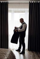 A man in a suit is standing in front of a window, holding a jacket. Concept of formality and professionalism, as the man is dressed in a suit and tie. The window provides natural light photo