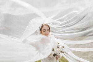 A bride is standing in front of a white veil, holding a bouquet of flowers. The scene is set in a field, with the bride's dress and veil creating a sense of elegance and romance photo
