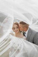 A bride and groom are kissing in front of a veil. The bride is wearing a white dress and the groom is wearing a gray suit. The scene is romantic and intimate photo