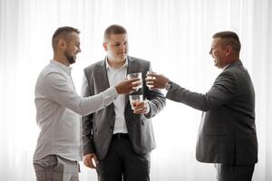 Three men are standing in a room, each holding a glass of liquor. They are all smiling and seem to be enjoying each other's company. Scene is lighthearted and social, as the men are sharing a drink photo