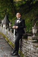 A man in a black suit and tie stands in front of a stone wall. He is wearing a white tie and a boutonniere photo