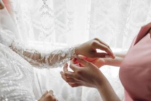 A woman is helping another woman put on a wedding dress. The scene is set in a room with a window photo