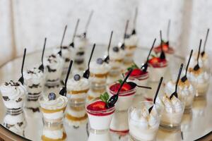 A table full of desserts with a variety of flavors and colors. The desserts are served in small plastic cups with spoons. The table is set up for a party or gathering photo