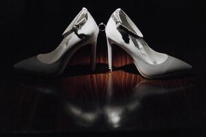 Two white high heels with a silver ring on the front. The shoes are on a wooden table photo