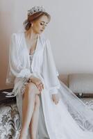A woman in a white gown sits on a bed. She is wearing a tiara and has her legs crossed photo