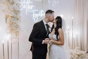 A bride and groom kiss in front of a white backdrop . The bride is holding a bouquet and the groom is holding a wine glass photo