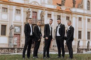 A group of five men are standing in front of a large building, dressed in black suits and ties. Scene is formal and elegant, as the men are posing for a photo in their suits