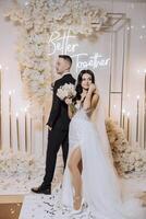 A bride and groom kiss in front of a white backdrop . The bride is holding a bouquet and the groom is holding a wine glass photo