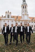 A group of five men in suits are walking together in front of a large building. The men are dressed in black and white, and they appear to be posing for a photo. Scene is formal and elegant photo