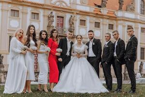 A group of people are posing for a picture in front of a building. The bride and groom are in the center of the group, with the bride wearing a white dress and the groom wearing a suit photo