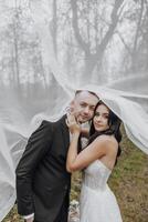 A man and woman are posing for a picture under a white veil. The man is wearing a suit and tie, while the woman is wearing a white dress. Scene is romantic and intimate photo